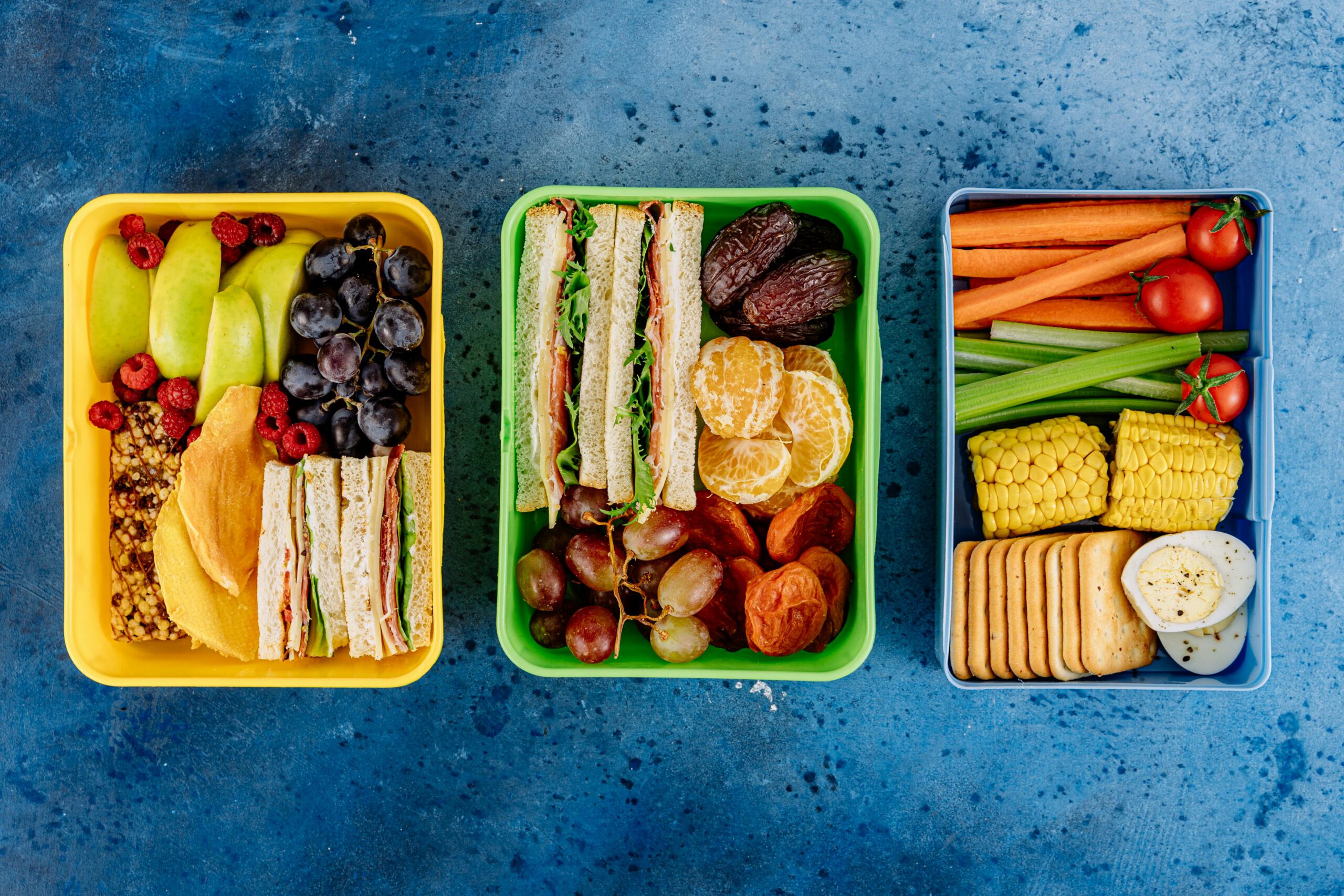 Three vibrant lunch boxes showcasing fruits, sandwiches, and vegetables on a blue textured background.
